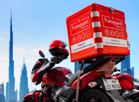 Red delivery scooter with a large red insulated box labeled 'Transguard Delivery' mounted on the back, parked against a city skyline of tall blue skyscrapers.